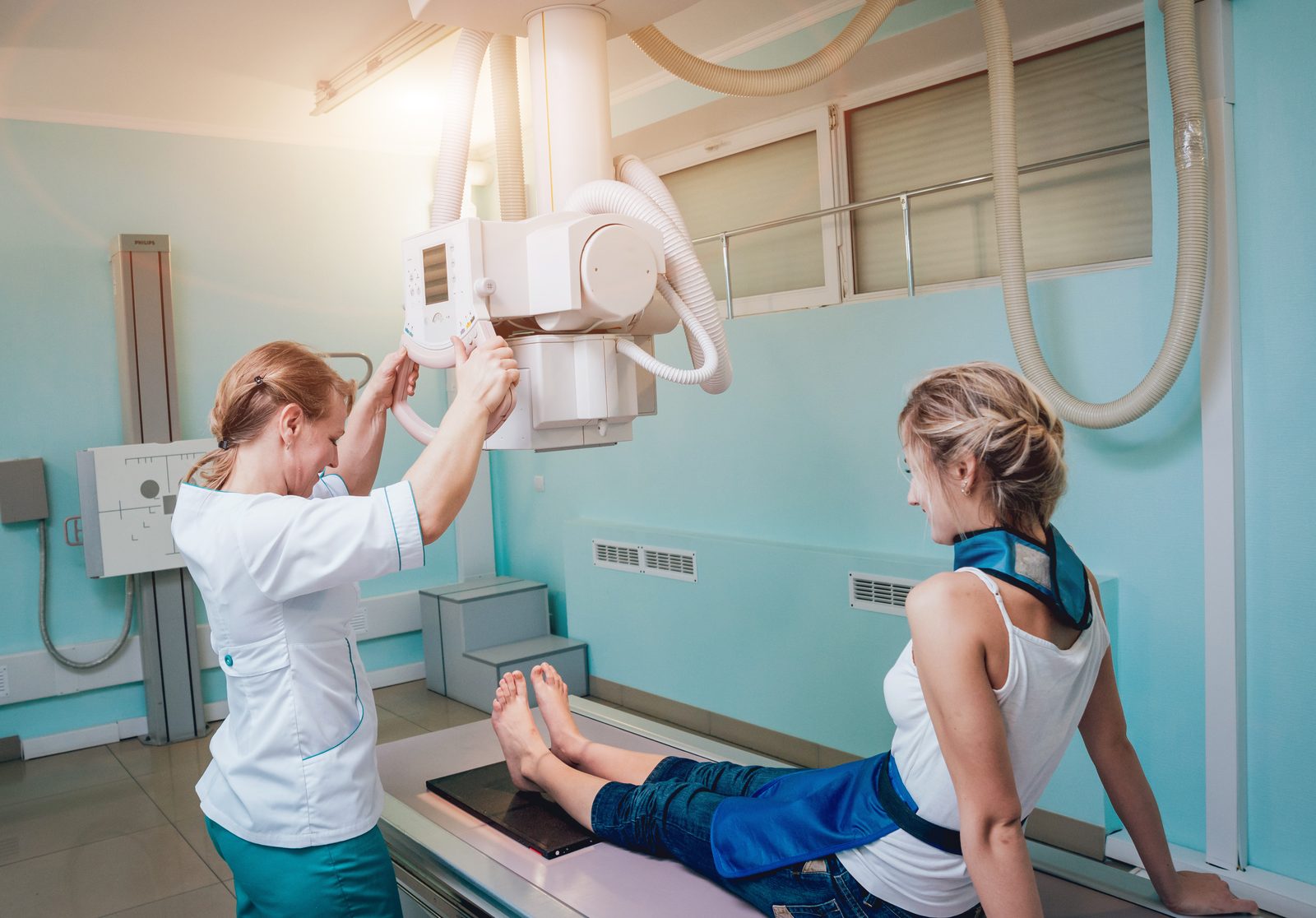 A technologist positioning the overhead X-ray tube for a patient wearing a lead apron.